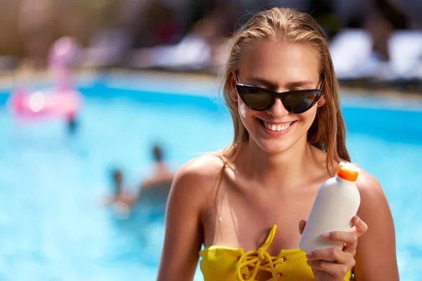 Blonde woman applies sunscreen solar cream on cheeck and face near swimming pool. Smiling pretty girl puts suntan cream from plastic bottle on skin in spa. Female in bikini with a suntan lotion.