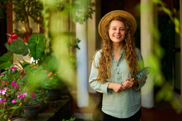 stock image Young woman gardener in straw hat holding garden scoop stands in garden looking at camera smiling happily. Cheerful caucasian female with orchard in the background. Gardening and farming concept