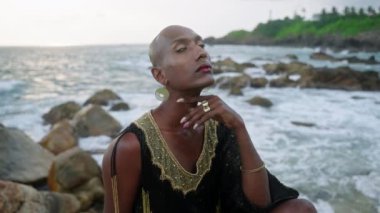 Renkli Androgynous taşın üzerinde oturuyor. Lüks elbiseli karamsar suratlı, kayalık okyanus sahillerinde takılar takıyor. Trans seks mankeni, burun halkası piercing, yüzük, küpe takıyor..