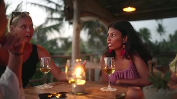 多様な女性の友人は日没時に屋上の海の景色レストランでワインを飲む 夕暮れ時に熱帯カフェのテーブルでキャンドルを語り 笑顔の多人種の女性 若い多民族女性の食事 — ストック動画