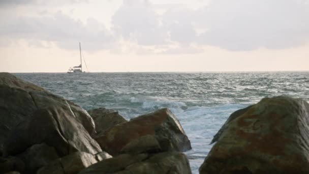 夕日の美しい海の景色でヨット船のセーリング 大きな岩やスプラッシュに対する波の休憩 岩のビーチで息をのむような海の夕日 ラフ水 瞑想の海を背景にした帆船 — ストック動画