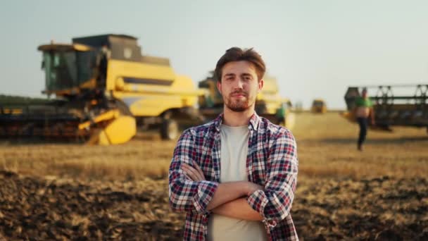 収穫した小麦畑に手を胸に交差して誇り高い農家の肖像画 ハーベスターマシンのドライバーが彼の組み合わせで立っている 笑みを浮かべて農学者のカメラを見ている 収穫作業の牧場主 — ストック動画