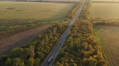 Yaz günbatımında güzel bir kırsalda, yolda giden tahıl kamyonunun hava görüntüsü. Kırsal alanda tarımsal alanlar ve ağaçlarla otoyol boyunca giden bir kamyonun insansız hava aracı görüntüsü.