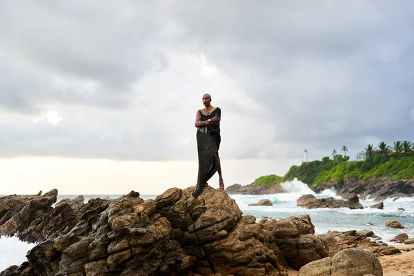 Non-binary black godlike person poses gracefully standing on rocks in ocean. Trans ethnic fashion model in a posh dress and jewellery on rocky beach by storm . Lgbtq. Pride month