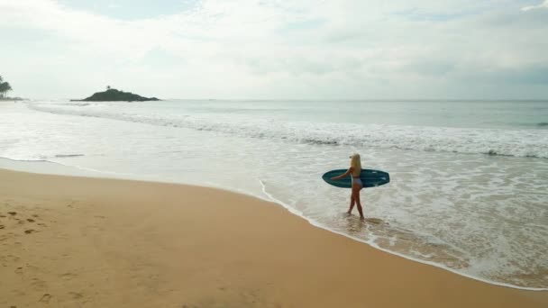 身穿泳衣的女冲浪者手持冲浪板在海滩上通过海空观看 穿着比基尼的年轻高加索女性从无人驾驶飞机的热带位置出发 沿着海滨的潮水带着冲浪板 — 图库视频影像
