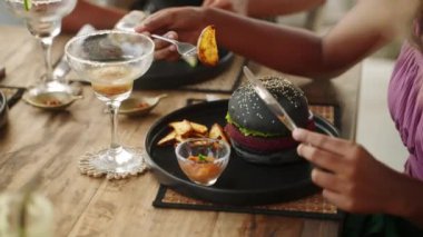 Restoranda hamburger ve patates kızartması yiyen zenci bir kadın. Bronzlaşmış kadın, otantik kafede akşam yemeği için patates dilimli siyah burger yiyor. Etnik kız iştah açıcı vejetaryen yemeğinin tadını çıkarıyor..