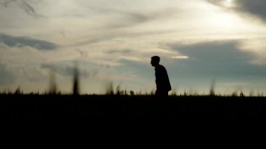 Çiftçi silueti çavdar tarlasında yürüyor ve olgunlaşmış buğday kulaklarına eliyle dokunuyor. Agronomist adam tahıl tarlasından manzaralı bir gökyüzüyle geçiyor. Tarım, hasat, tarım ve gıda tedarik kavramı.