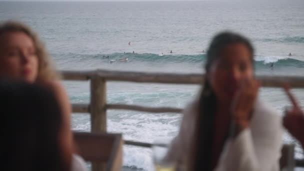 アウトドアシービューレストランでは 様々な女性の友人は ラインナップにサーファーを見て海を見ている 熱帯の海の見えるカフェのテラスのテーブルに座っている多人種の女の子 ゴシップ — ストック動画