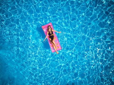 Yüzme havuzunda bikinili güzel bir kız şişme pembe yatakta dinleniyor. Mayo bronzlaştırıcı giymiş zayıf seksi bir kadın. Lüks oteldeki mavi suda dinlenen bir kadın. Gökyüzü, yukarıdan görünüm.