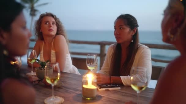 多様な女性の友人は日没時に屋上の海の景色レストランでワインを飲む 夕暮れ時に熱帯カフェのテーブルでキャンドルを語り 笑顔の多人種の女性 若い多民族女性の食事 — ストック動画