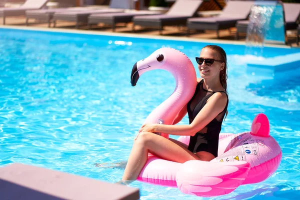Hot slim woman having fun laughing on inflatable pink flamingo float mattress in bikini at swimming pool. Pretty female on tropical vacation. Attractive fit girl in swimwear lies in the sun on floaty.
