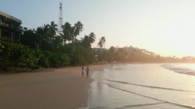 Genç, çok ırklı bir çift okyanus kıyısındaki tropikal bir sahilde gün doğumunda drone görüntülerinde koşuyor. Erkek arkadaşı ve kız arkadaşı şafak vakti deniz kenarında deniz kenarında su oyunu etiketinin yanında koşuyorlar.