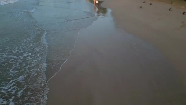 日の出に砂の上を走る波 若いカップルは 手空の景色を保持する潮に歩いています ボーイフレンドとガールフレンドが行く ドローンからの夜明けのバックビュー映像で海辺でビーチで話す — ストック動画
