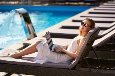 Güzel sarışın kadın, spa 'daki havuz yatağında dizüstü bilgisayarla uzaktan kumanda yapıyor. Serbest çalışan iş kadını, tropikal ada tatil köyünden bir ekiple telekomünikasyon yapıyor. Kız güneşleniyor ve dinleniyor..