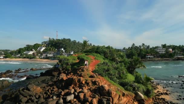 波の空中ビューで洗浄海の真ん中に緑の熱帯岩の島の上にカップル立っている スリランカのミリッサにある有名なオウム岩は ドローンから海の映像に囲まれています — ストック動画