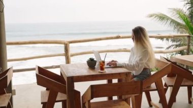 Kafede dizüstü bilgisayarla çalışan, okyanus manzaralı, palmiye ağaçları olan bir kadın. Lüks bir restoranda oturan gezgin blog yazarları. Uzaktan çalışma gününde SMM yöneticisi. Kadın deniz kenarında buzlu kahve içiyor..