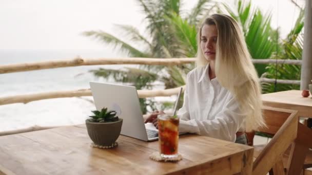 オーシャンビューのヤシの木とカフェでラップトップで作業するかなり女性 トロピカルレストランに座っているコンピュータのキーボードをタイプする女性 リモートワーキングデーのビジネスマン 素敵な女の子は冷たいお茶を飲む — ストック動画