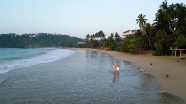 Güneşin doğuşunda deniz boyunca tropikal sahilde el ele tutuşan çift hava aracı görüntülerinde yürüyor. Hindistan cevizli erkek arkadaş ve kız arkadaş şafak vakti deniz kenarında deniz kenarında yürüyorlar.