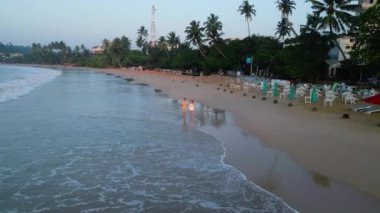 Güneşin doğuşunda deniz boyunca tropikal sahilde el ele tutuşan çift hava aracı görüntülerinde yürüyor. Hindistan cevizli erkek arkadaş ve kız arkadaş şafak vakti deniz kenarında deniz kenarında yürüyorlar.