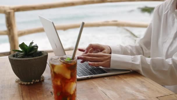 オーシャンビューの屋外トロピカルカフェのラップトップキーボードに入力する女性 アイスティーで屋外のコワーキングに座っているノートを使用してテキストメッセージング リモートワークデーのビジネスマンと飲み物 — ストック動画