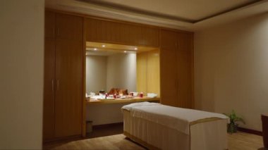 Geniş bir spa odasının ortasında masaj yatağı. Arka planda büyük bir ayna ve mumlar var. Loş ışık, beyaz battaniye ve havlularla kaplı masaj masası olan bir sağlık merkezi süiti.