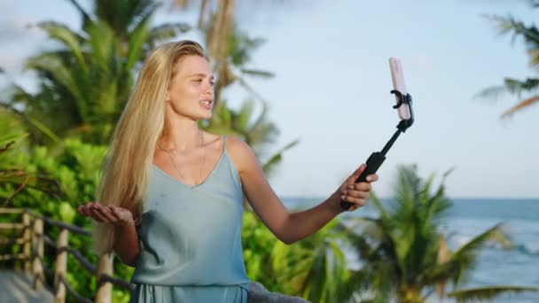 ブロンドの女性の側面のビューは 海を望む熱帯島のセルフィーカメラで自分自身を撮影するブロガーを旅行します 女性のVooggerはスリランカのブログのための電話と記録ビデオが付いているセルフィースティックを保持します — ストック動画