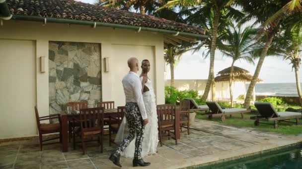 婚礼结束后 已婚男性穿着时髦的婚纱和西服 穿着热带岛屿上的游泳池在别墅里散步 一对同性恋夫妇在海边的度假胜地和异国情调的棕榈树旁度蜜月 — 图库视频影像