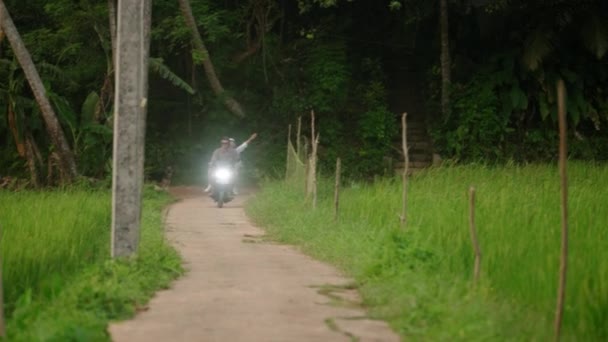 男人骑摩托车 女人笑 在热带地区骑摩托车 恋爱中的夫妻驾驶摩托车沿着稻田在丛林的路上 骑自行车旅行的愉快游客 度蜜月 — 图库视频影像