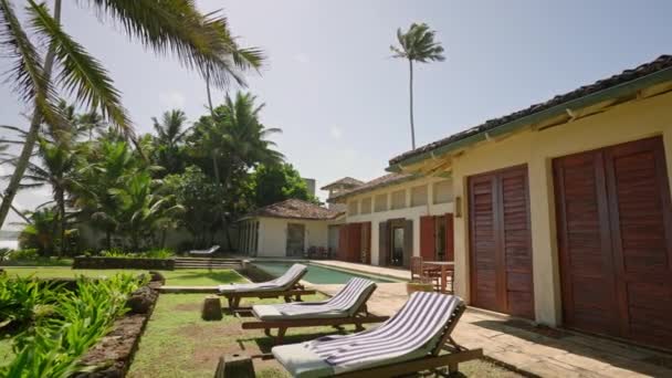热带岛屿沙滩海岸附近的豪华度假别墅 有游泳池和绿草 还有棕榈树 蓝色的温水 有涟漪 有舒适的院子 还有供游客休息的太阳 — 图库视频影像