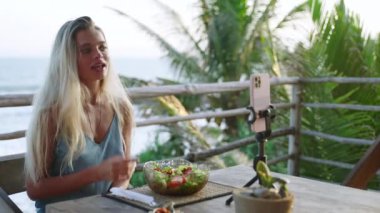 Yemek blogcusu kadın yeşil salata kasesiyle ilgili videolar çekiyor, kafede sıska bir vücut için diyet yapıyor, deniz manzaralı. Seyahat etmenleri yerel mutfağı, sosyal ağdaki diyeti anlatır. Vejetaryen kadın bir vlog çekiyor.