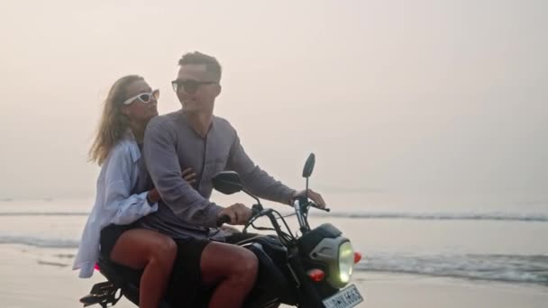 日出时 一对旅行的夫妇骑摩托车沿着沙滩行驶 女性游客在海洋海岸线上驾驶租来的摩托车 女士拥抱男人 张开双臂 笑着庆祝骑自行车的自由 — 图库视频影像