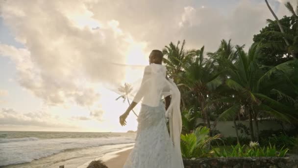 白いシフォンのウェディングドレスで海岸にポーズするフランボイアントジェンダー流体黒人 カメラを見る非バイナリーの花嫁は 熱帯の島の海浜で日没を楽しんでいます ビポックアンドロマンスのファッションモデル — ストック動画