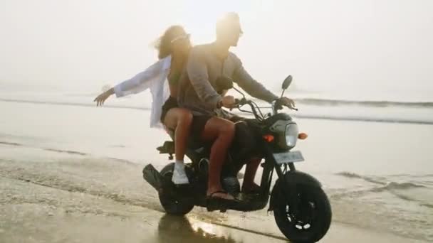 日出时 快乐的人们骑摩托车在海滨泼水 恋爱中的夫妻在海滨驾驶摩托车很开心 女孩拥抱男朋友 太阳透镜耀斑 慢动作 — 图库视频影像