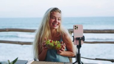 Kadın kendini tropikal adada yemek yerken videoya çekiyor. Kadın blogcu yeşil sağlıklı salata bolyasını tutuyor ve kamerasına gülümsüyor. Açık kafedeki okyanus manzaralı tripodun üzerinde duruyor..