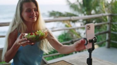 Yemek blogcusu kadın yeşil salata kasesiyle ilgili videolar çekiyor, kafede sıska bir vücut için diyet yapıyor, deniz manzaralı. Seyahat etmenleri yerel mutfağı, sosyal ağdaki diyeti anlatır. Vejetaryen kadın bir vlog çekiyor.