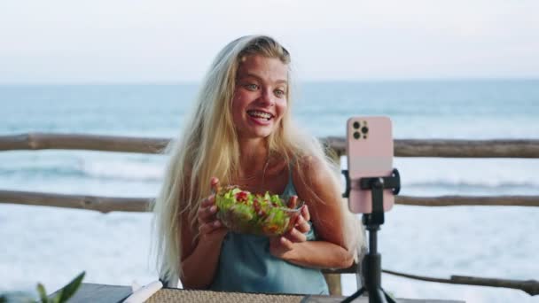 女人拍摄她自己在热带岛屿上吃午饭的录像 女性博主拿着绿色健康的沙拉 站在三脚架上的相机上微笑着 在户外咖啡馆看到大海 — 图库视频影像