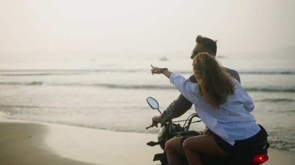 日出时分 一对快乐的夫妇骑摩托车在海滨 女人驾驶摩托车在海岸 穿着比基尼 太阳镜的漂亮女士回头看相机 风吹着她的头发 — 图库视频影像
