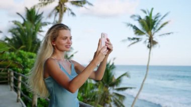 Kadın telefonunda cennet manzarası, okyanus, gökyüzü ve palmiye ağaçlarının videosunu çekiyor. Kız seyahat blogcusu sıcak yaz tatillerinde açık hava kafesindeki tropikal adanın manzarasını filme alır..