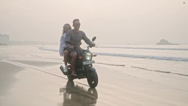日出时分 一对旅行的夫妇骑摩托车在潮湿的沙滩上 女人驾驶租来的滑板车沿着海岸线的海洋 女士拥抱着男朋友 张开双臂迎风 庆祝自由 — 图库视频影像