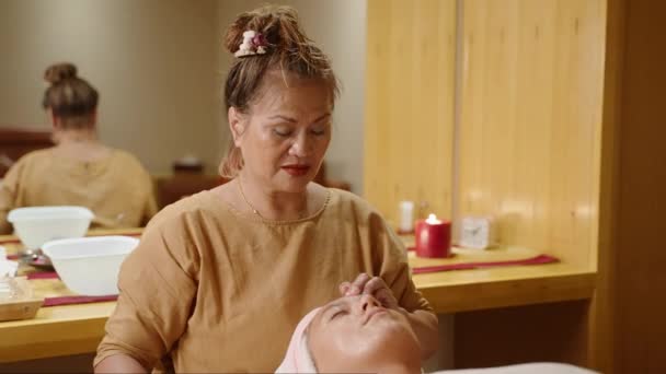 年长的亚洲按摩师用双手按摩年轻的女客户 在温泉中使用油 健康中心的成年女工给一个涂满白毛巾的晒黑的女孩做了放松的面部按摩 — 图库视频影像