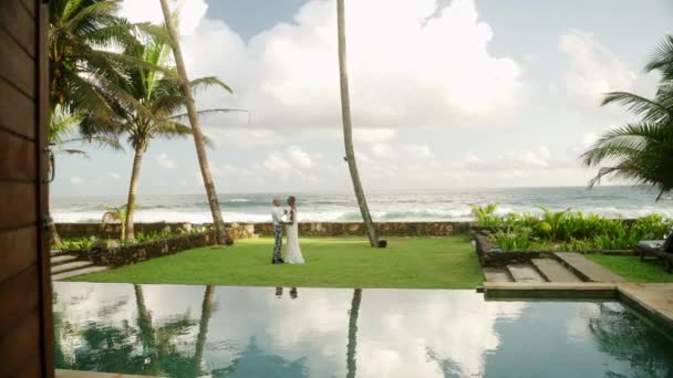 新结婚的一对同性恋夫妇在草坪上跳舞 在海边的豪华复古别墅游泳池边跳舞 在热带地区的婚礼上 穿着婚纱和白衬衫的时髦新婚夫妇拥抱在一起 — 图库视频影像