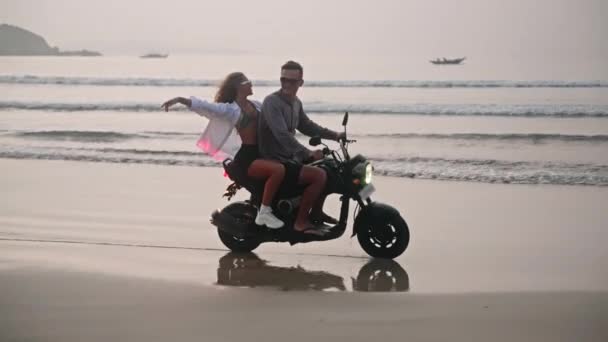 浪漫的夫妻在日出时分骑摩托车在海滩上约会 情侣们在海岸线上骑摩托车旅行 带着倒影和水花 女朋友抱着她的男朋友 张开双臂 — 图库视频影像