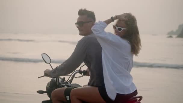 一对年轻夫妇在日出时骑摩托车在海滩上 女人骑摩托车沿着海岸线在海上旅行 女士转过头对着相机笑着张开胳膊在风中吹拂着头发 — 图库视频影像