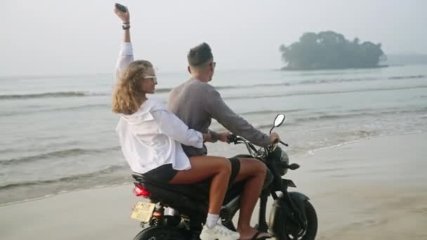 日出时年轻人在海滨骑摩托车 女人驾驶摩托车在海岸 穿着比基尼 太阳镜的漂亮姑娘回头看相机 笑着举手 风吹头发 — 图库视频影像