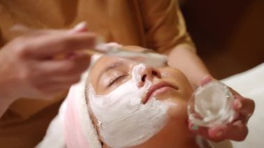 Güzellik uzmanı, kadın hasta yüzüne sağlık merkezinde bir fırçayla beyaz kilden maske takıyor. Kozmetik uzmanı, kaplıca çekiminde genç bir kadına yüz bakımı prosedürü uyguluyor. Güzellik ve yüz bakımı kavramı