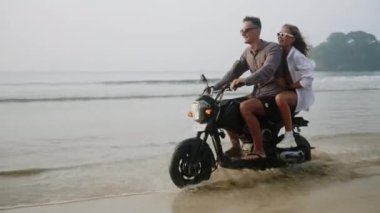 Genç çift gün batımında ıslak kumlu sahilde su sıçratarak motosiklet sürüyor. Dostum, kadınlar okyanus kıyısında motosiklet sürerken eğleniyor. Kadın bağırıyor, erkek arkadaşına sarılıyor ve romantik bir yolculuğa çıkıyor. Yavaş çekim.