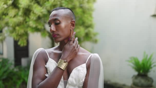 ゲイ黒人男性はウェディングドレス 真鍮のイヤリング ジュエリーでポーズします 非伝統的な花嫁は美しさ きちんとしたマニキュア メイクアップ 宝石のブレスレットを示しています ヴィラグリーンガーデンでのLgbtqファッション撮影 — ストック動画