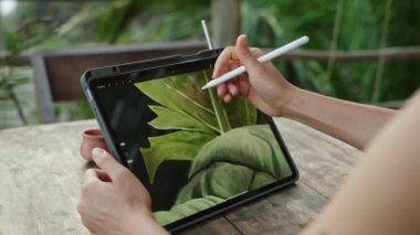 Kadın sanatçı elleri, Stylus 'la birlikte tropik adadaki kafede, palmiye ağaçlarında resim çiziyor. Genç tasarımcı egzotik mekanlarda açık havada çalışıyor. İllüstrasyon düzenleyen güzel bir kadın. Yakın plan..