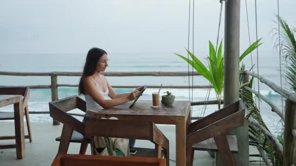 在海滨餐厅 棕榈树餐厅工作的女自由职业者 年轻女子编辑平板电脑上的插图 在海滨咖啡店喝冰咖啡 在偏远工作日工作的商人 — 图库视频影像