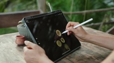 Kadın sanatçı elleri, Stylus 'la birlikte tropik adadaki kafede, palmiye ağaçlarında resim çiziyor. Genç tasarımcı egzotik mekanlarda açık havada çalışıyor. İllüstrasyon düzenleyen güzel bir kadın. Yakın plan..